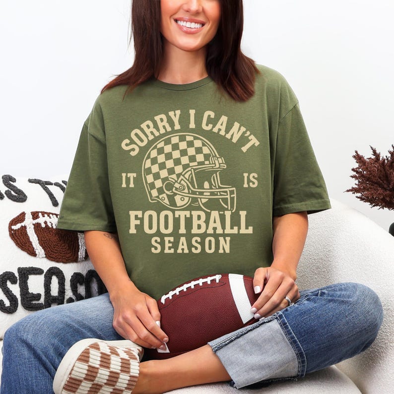 K&ouml;nnte beinhalten: Olivgr&uuml;nes T-Shirt mit dem Text "SORRY I CAN'T IT IS FOOTBALL SEASON" und einer Footballhelm-Grafik. Die Person h&auml;lt einen braunen und wei&szlig;en Football. Das Shirt wird mit Jeans und braunen und wei&szlig;en Schuhen kombiniert.
