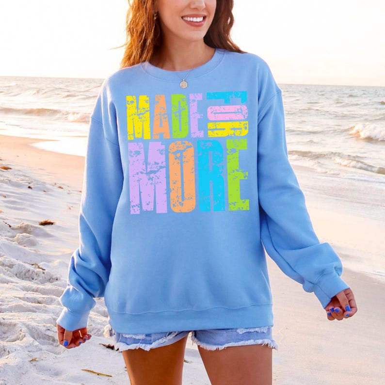 May include: Light blue sweatshirt with the words "MADE MORE" in large, colorful, distressed text. The text is in yellow, pink, purple, orange, and green. The sweatshirt has long sleeves and is being worn on a beach.
