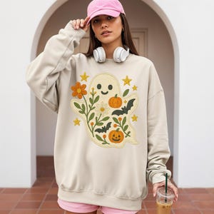 May include: Beige sweatshirt with a ghost design, pumpkins, flowers, bats, and stars. The ghost is surrounded by orange, green, and white floral elements. The sweatshirt has long sleeves and a crew neck.