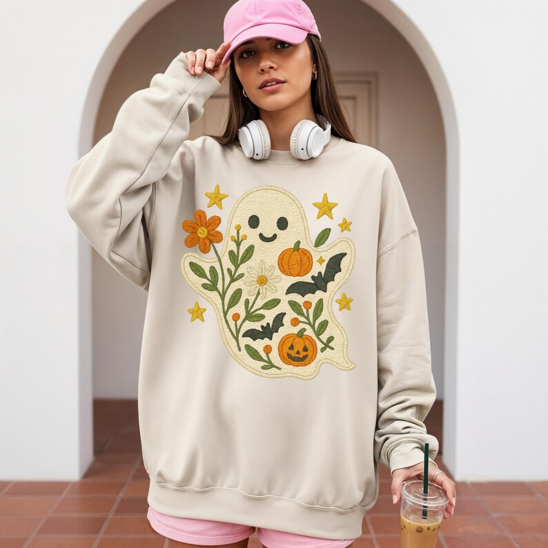 May include: Beige sweatshirt with a ghost design, pumpkins, flowers, bats, and stars. The ghost is surrounded by orange, green, and white floral elements. The sweatshirt has long sleeves and a crew neck.
