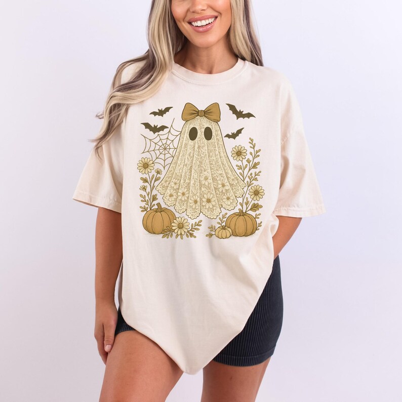 Puede incluir: Camiseta color crema con un dise&ntilde;o de Halloween. El dise&ntilde;o incluye un fantasma con un lazo, calabazas, murci&eacute;lagos y una telara&ntilde;a, rodeado de flores. La camiseta tiene mangas cortas y un corte holgado.