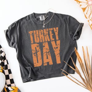 May include: A dark gray t-shirt with the words "TURKEY DAY" printed in orange, distressed lettering. The shirt has rolled-up sleeves and a crew neck. A silver necklace with a pendant is draped on the shirt.