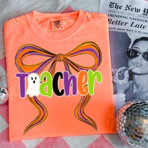 May include: A neon orange t-shirt with a large bow design in orange, purple, and black. The word "Teacher" is printed across the shirt in purple, green, black, and orange, with a ghost graphic. The shirt is a Comfort Colors brand.