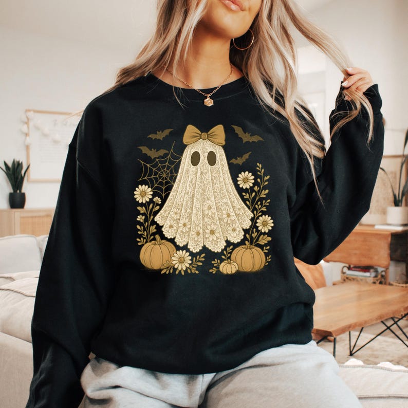 Puede incluir: Sudadera negra con un dise&ntilde;o de fantasma beige con lazo, rodeado de calabazas, flores, murci&eacute;lagos y una telara&ntilde;a. El fantasma tiene dos ojos ovalados y lleva un lazo. El dise&ntilde;o es de estilo vintage, perfecto para Halloween.