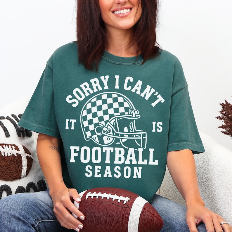 K&ouml;nnte beinhalten: Dunkelgr&uuml;nes T-Shirt mit dem wei&szlig;en Text "SORRY I CAN'T IT IS FOOTBALL SEASON". Das Shirt zeigt eine Footballhelm-Grafik mit Schachbrettmuster. Ein brauner und wei&szlig;er Football wird im Scho&szlig; gehalten.