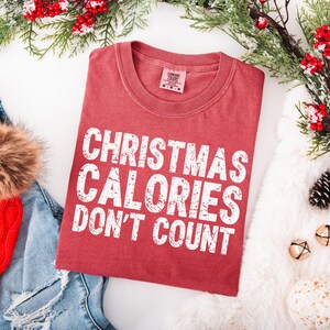 May include: A red t-shirt with the text "CHRISTMAS CALORIES DON'T COUNT" in white block letters. The shirt is folded and surrounded by holiday decorations, including greenery and a red hat.
