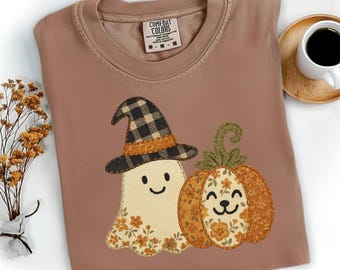 May include: A brown Comfort Colors t-shirt featuring a Halloween-themed design. The design includes a smiling ghost wearing a plaid witch hat and a smiling pumpkin. A cup of coffee sits on a wooden coaster.