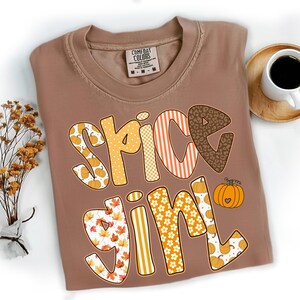 Puede incluir: Una camiseta marrón con las palabras "Spice Girl" en un diseño colorido y estampado. Las letras presentan calabazas, hojas, rayas y estampado de leopardo. Un pequeño gráfico de calabaza está al lado del texto. La camiseta está al lado de una taza de café.