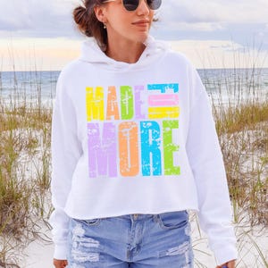 May include: White cropped hoodie with the words "MADE MORE" in colorful, distressed lettering. The text is in yellow, green, pink, purple, and orange. The hoodie is paired with blue denim shorts.