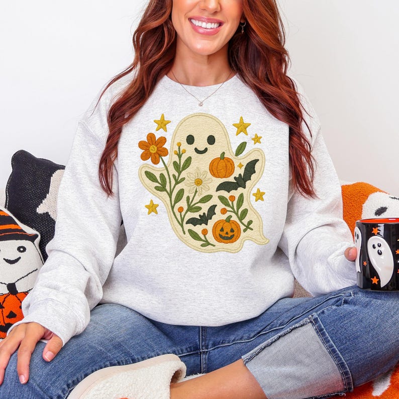 May include: A light grey sweatshirt with a whimsical ghost design. The ghost is decorated with orange pumpkins, flowers, and bats, with yellow stars scattered around. The design has a cheerful, autumnal theme.