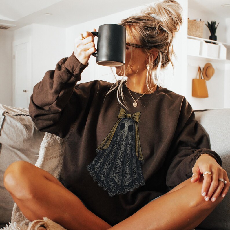 K&ouml;nnte beinhalten: Ein braunes Sweatshirt mit einem Geisterdesign. Der Geist hat eine Schleife und ist in schwarzer Spitze drapiert. Das Sweatshirt ist einfarbig braun und hat lange &Auml;rmel. Eine schwarze Tasse wird gehalten.