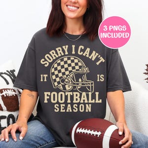 K&ouml;nnte beinhalten: Dunkelgraues T-Shirt mit dem Text "SORRY I CAN'T IT IS FOOTBALL SEASON" und einer Footballhelm-Grafik. Ein rosa Kreis oben rechts zeigt "3 PNGS INCLUDED". Ein Football wird im Scho&szlig; gehalten.