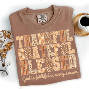 以下が含まれることがあります： 茶色のComfort Colors Tシャツ。パッチワークデザインで「THANKFUL, GRATEFUL, BLESSED」の文字がプリントされています。「God is faithful in every season」の文字が下にあります。背景にはコーヒーカップとドライフラワーがあります。