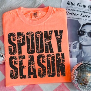 May include: Bright orange t-shirt with the words "SPOOKY SEASON" in large, distressed black letters. The shirt is a crew neck and appears to be made of a soft material. A newspaper with the headline "Better Late" is in the background.