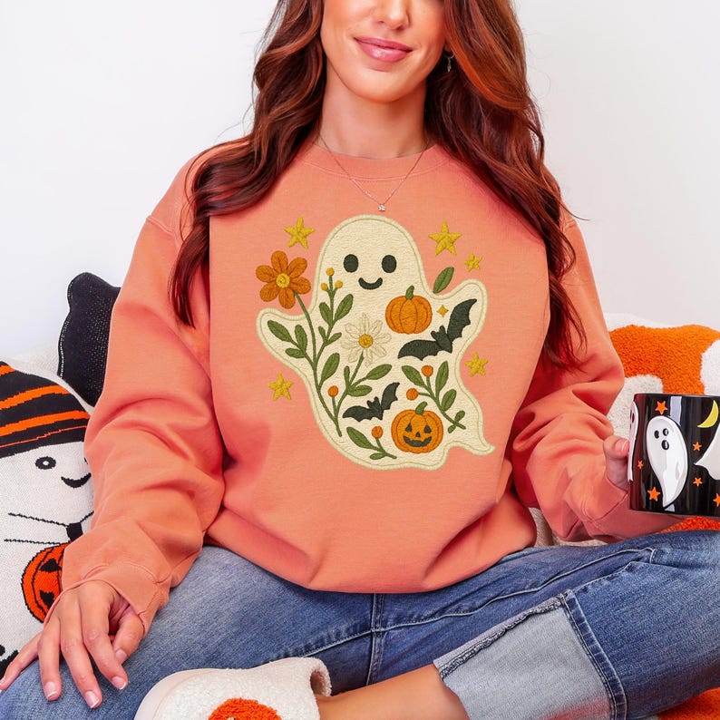 May include: Coral-coloured sweatshirt with an embroidered ghost design, including a smiling face, flowers, pumpkins, bats, and stars. The sweatshirt is worn with blue jeans. A Halloween-themed mug is also visible.