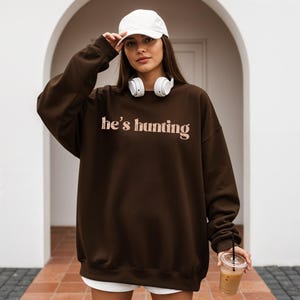 May include: A dark brown sweatshirt with the text "he's hunting" in a light pink font. The sweatshirt is worn with a white baseball cap and white headphones. The person is holding a clear plastic cup with a straw.