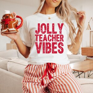 May include: White t-shirt with the text "JOLLY TEACHER VIBES" in red. The person is wearing red and white striped pajama pants and holding a gingerbread man mug. The mug has a red handle and a red scarf.