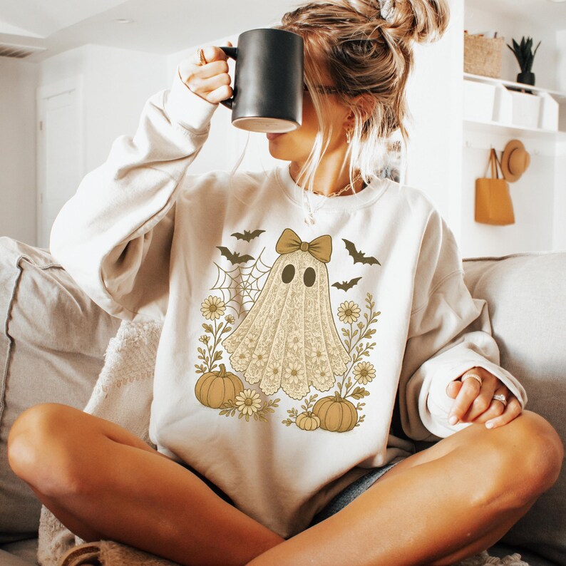 Puede incluir: Sudadera color crema con un dise&ntilde;o de fantasma con un lazo, rodeado de calabazas, flores, murci&eacute;lagos y una telara&ntilde;a. El fantasma es de encaje. Una taza negra se sostiene frente a la cara.