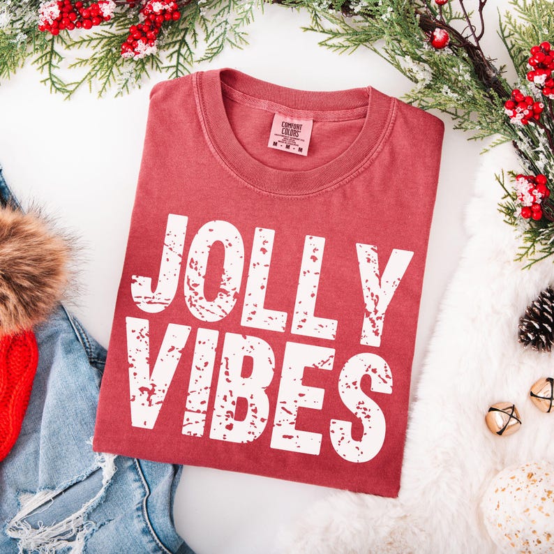 May include: A red t-shirt with the words "JOLLY VIBES" in white, distressed lettering. The shirt is folded and surrounded by holiday decorations, including greenery, berries, and a white fluffy surface. A red knit hat and denim jeans are visible.