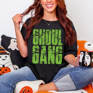 May include: Black t-shirt with the text "GHOUL GANG" in distressed green lettering. The person is wearing blue jeans and holding a black mug with white ghost designs. The theme is Halloween.