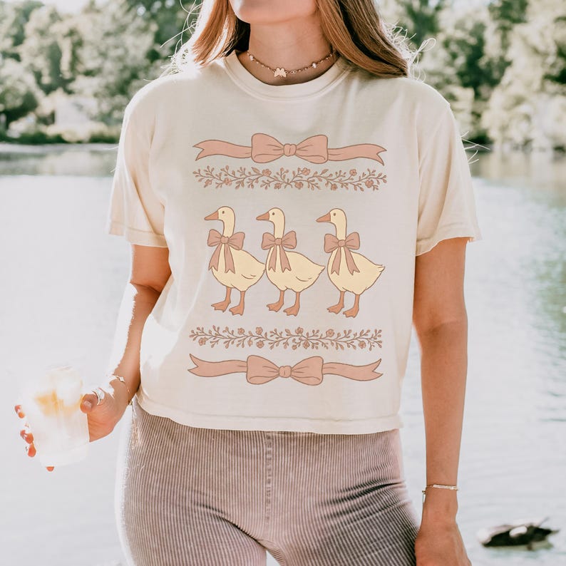 May include: A white cropped t-shirt with a graphic of three ducks wearing pink bows. The ducks are in a line and are facing the right. The graphic is surrounded by a floral design and pink ribbons with bows.