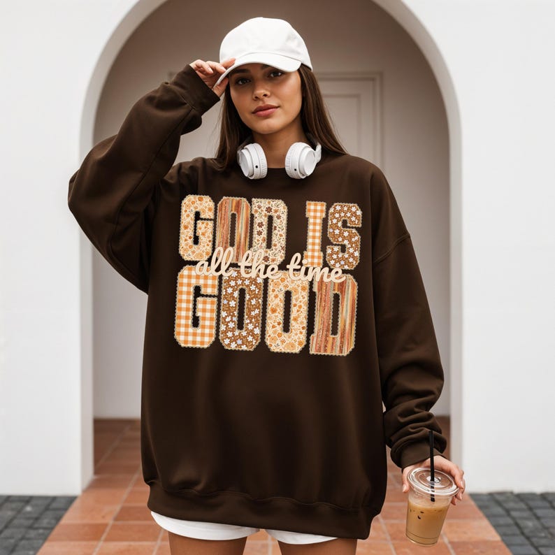 May include: A dark brown sweatshirt with the text "GOD IS all the time GOOD" in a mix of patterned fonts. The patterns include floral, plaid, and wood grain designs. The person in the image is wearing a white baseball cap and headphones.