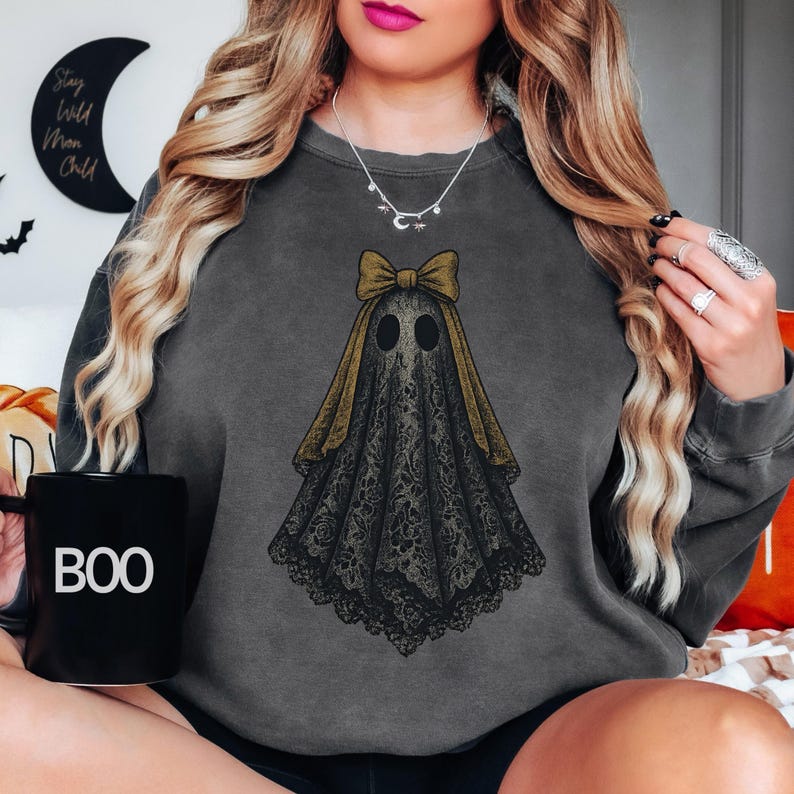 K&ouml;nnte beinhalten: Dunkelgraues Sweatshirt mit einem Geisterdesign, einer goldenen Schleife und Spitzendetails. Der Geist hat zwei schwarze ovale Augen. Eine schwarze Tasse mit dem Wort "BOO" ist zu sehen. Halloween-Kleidung.
