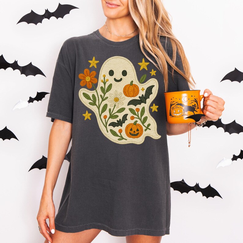 May include: Dark grey t-shirt with a friendly ghost design, pumpkins, flowers, and bats. The ghost is surrounded by floral elements and stars. An orange mug with a Halloween scene is held in the hand.