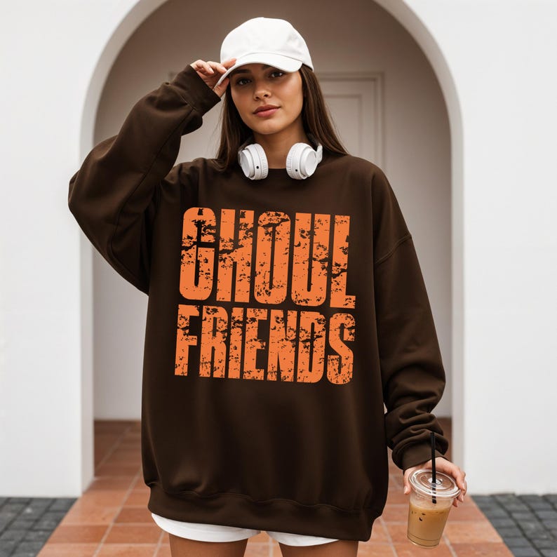 May include: A brown sweatshirt with the text "GHOUL FRIENDS" in orange. The sweatshirt is worn by a person wearing a white baseball cap and white headphones. The person is holding a clear plastic cup with a straw.