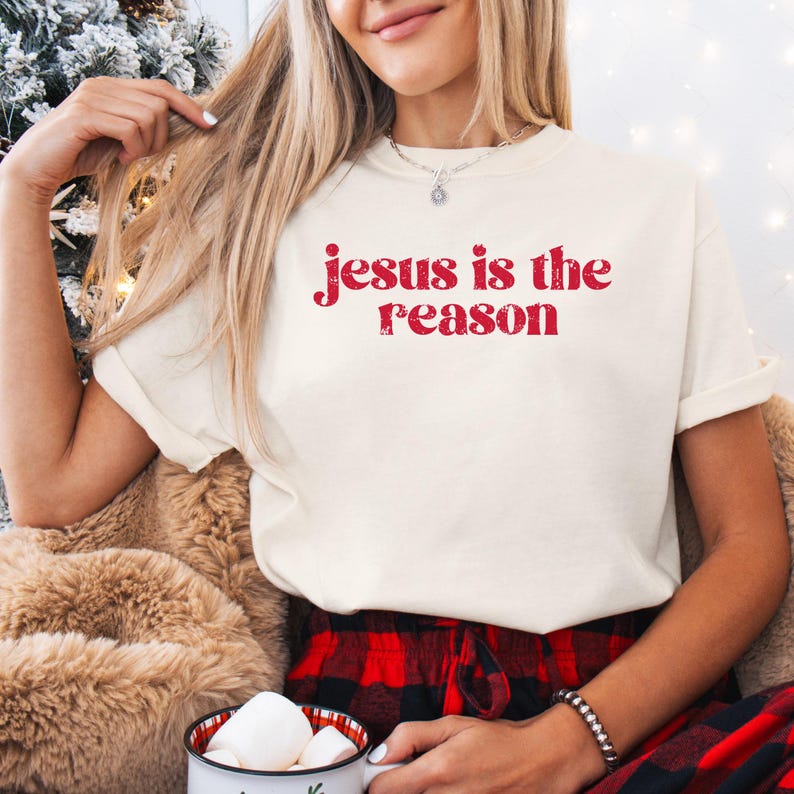 Puede incluir: Camiseta color crema con el texto rojo "Jesus is the reason". La camiseta tiene las mangas dobladas. La persona lleva pantalones de cuadros rojos y negros y sostiene una taza con malvaviscos.