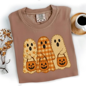 May include: A brown Comfort Colors t-shirt featuring three ghost characters with jack-o'-lantern treat bags. The ghosts have different patterns: floral, plaid, and solid. A cup of coffee sits on a wooden coaster.