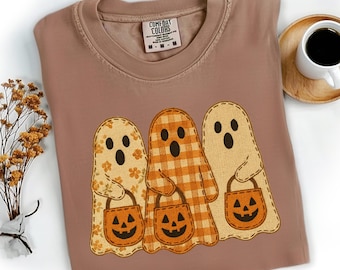 May include: A brown Comfort Colors t-shirt featuring three ghost characters with jack-o'-lantern treat bags. The ghosts have different patterns: floral, plaid, and solid. A cup of coffee sits on a wooden coaster.