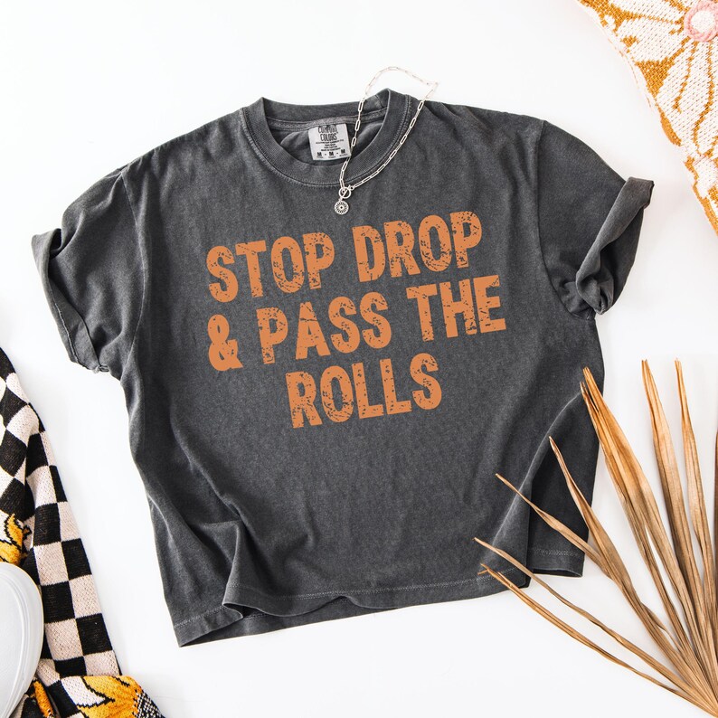 Pu&ograve; includere: T-shirt grigio scuro con la scritta arancione "STOP DROP & PASS THE ROLLS". La maglietta ha un girocollo e maniche corte. Una collana d'argento con un ciondolo &egrave; drappeggiata sulla maglietta.
