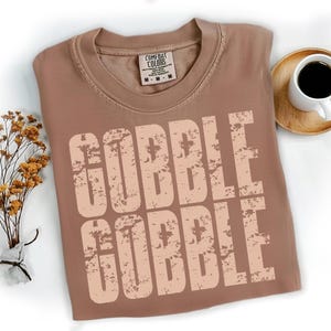 May include: A brown Comfort Colors t-shirt with the words "GOBBLE GOBBLE" printed in a distressed, light brown font. The shirt is folded and placed on a white surface, with a small bouquet of dried flowers and a cup of coffee nearby.