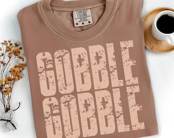 May include: A brown Comfort Colors t-shirt with the words "GOBBLE GOBBLE" printed in a distressed, light brown font. The shirt is folded and placed on a white surface, with a small bouquet of dried flowers and a cup of coffee nearby.