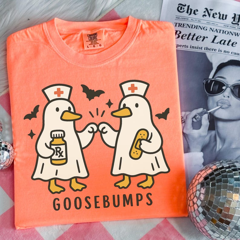 May include: Coral Comfort Colors t-shirt featuring a cartoon design of two goose characters dressed as nurses, with bats and stars. One goose holds a prescription bottle, the other a bandage. The word "GOOSEBUMPS" is printed below.