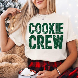 May include: Cream-colored t-shirt with the words "COOKIE CREW" in distressed green lettering. The shirt is worn with red and black plaid pants. A mug with marshmallows is held in the person's hands.