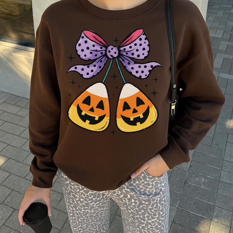 May include: A brown sweatshirt featuring a Halloween-themed graphic. The graphic includes two candy corn pieces with jack-o'-lantern faces, connected by a purple polka-dot bow with pink accents. The sweatshirt is long-sleeved.