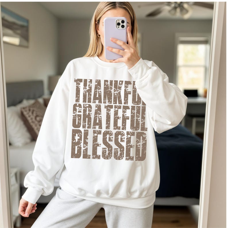 May include: White sweatshirt with the words "THANKFUL, GRATEFUL, BLESSED" in a distressed brown font. The sweatshirt has long sleeves and a crew neck. The image is taken in front of a mirror.