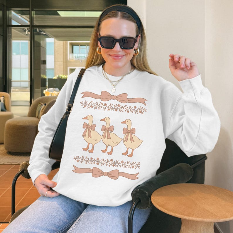 May include: White sweatshirt with a graphic of three ducks wearing bows. The ducks are in a line and have floral designs around them. The sweatshirt has a brown ribbon bow design at the bottom.