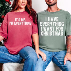 May include: Two people wearing matching t-shirts. One shirt is a dusty rose color and says "IT'S ME. I'M EVERYTHING" in white text. The other shirt is sage green and says "I HAVE EVERYTHING I WANT FOR CHRISTMAS" in white text. Both are wearing blue jeans.
