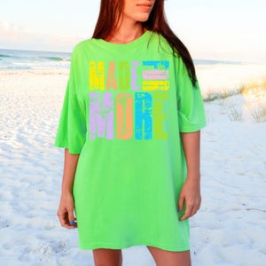 May include: A neon green t-shirt with the words "MADE FOR MORE" printed in colorful, distressed block letters. The shirt has short sleeves and a relaxed fit. The background is a sandy beach with the ocean in the distance.