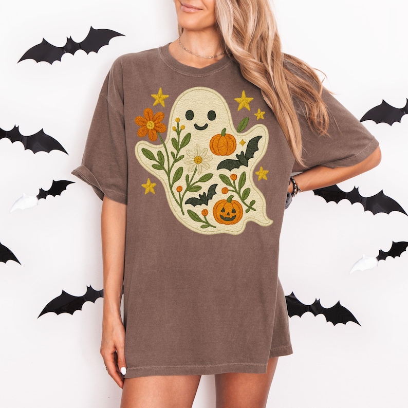 May include: Brown t-shirt with a ghost design, pumpkins, bats, flowers, and stars. The ghost has a smiling face and is surrounded by floral elements. Halloween-themed clothing.