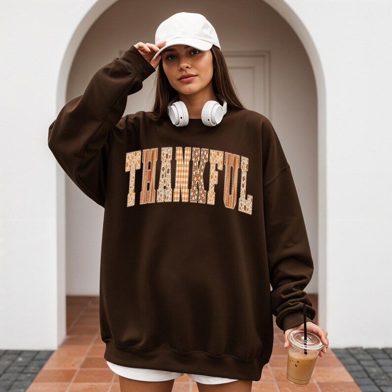 May include: A brown sweatshirt with the word "THANKFUL" in a patchwork design across the chest. The sweatshirt is worn with a white baseball cap and headphones. The person is holding a clear cup with a brown beverage.