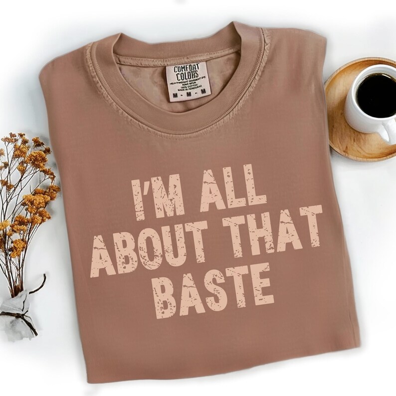 Pu&ograve; includere: Una maglietta marrone Comfort Colors con la scritta "I'M ALL ABOUT THAT BASTE" in un font invecchiato. La maglietta &egrave; piegata e posizionata su una superficie bianca, con un piccolo bouquet di fiori secchi e una tazza di caff&egrave; nelle vicinanze.