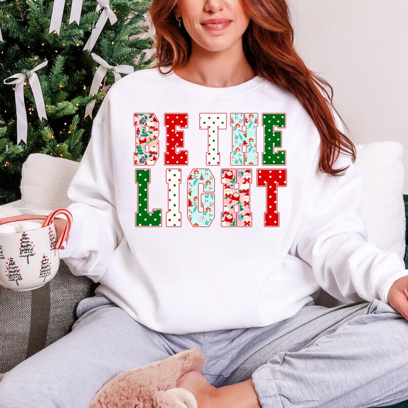 May include: White sweatshirt with the text "BE THE LIGHT" in red, green, and blue letters. The letters are decorated with Christmas-themed patterns. The person is sitting in front of a Christmas tree and holding a mug.