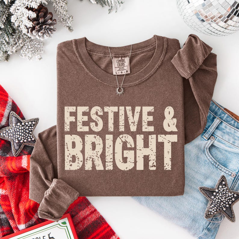 May include: A brown long-sleeve shirt with the words "FESTIVE & BRIGHT" in a distressed, off-white font. The shirt is paired with blue jeans and a silver necklace. Decorative stars and a disco ball are also in the image.