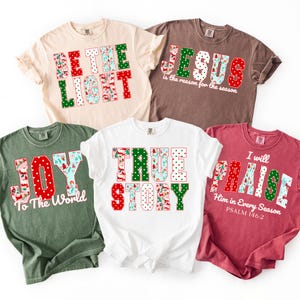 May include: Five t-shirts with Christmas-themed text designs. The shirts are in various colors, including beige, brown, green, white, and red. The text includes phrases like "BE THE LIGHT," "JESUS," "JOY," "TRUE STORY," and "PRAISE."