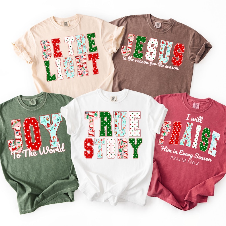 May include: Five t-shirts with Christmas-themed text designs. The shirts are in various colors, including beige, brown, green, white, and red. The text includes phrases like "BE THE LIGHT," "JESUS," "JOY," "TRUE STORY," and "PRAISE."
