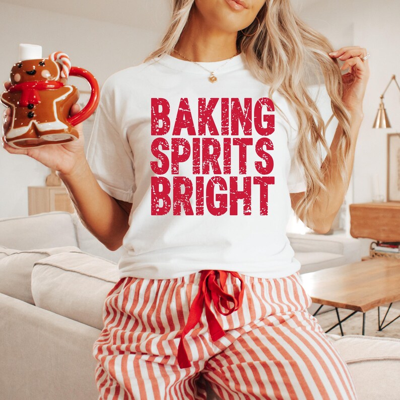 May include: A white t-shirt with the text "BAKING SPIRITS BRIGHT" in red letters. The person is wearing red and white striped pajama pants and holding a gingerbread man mug with a red handle.